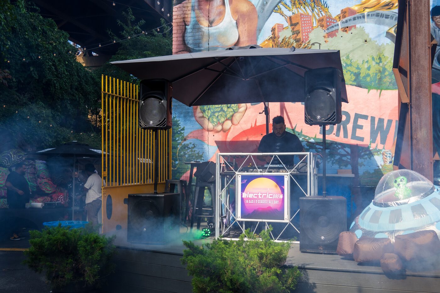 A DJ performing at Electric Brew, a day-into-night Sirens & Kings event, framed by speakers and a colorful Bronx mural.