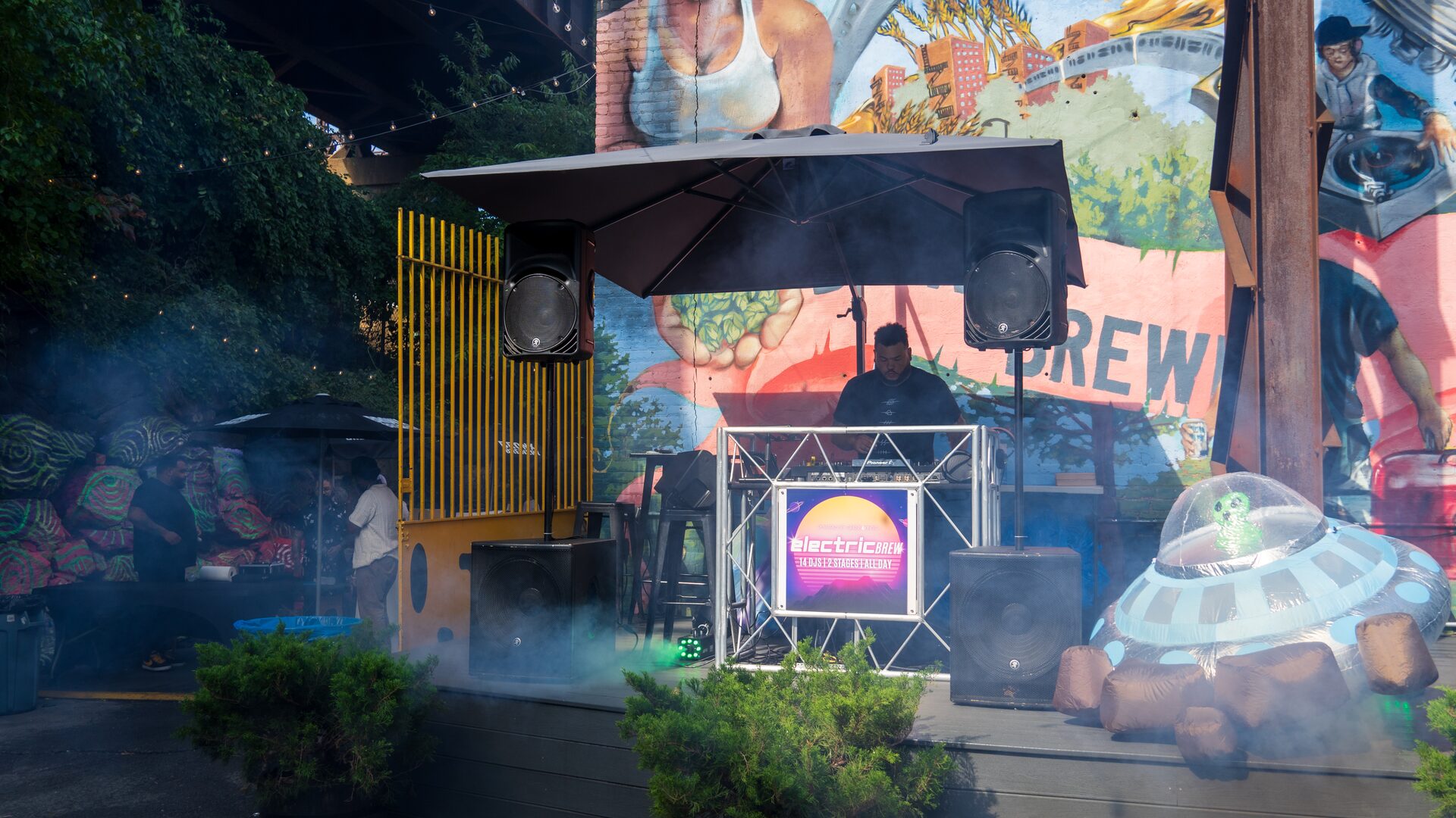 A DJ performing at Electric Brew, a Sirens & Kings event, with stage speakers, haze and a Bronx mural backdrop at dusk.